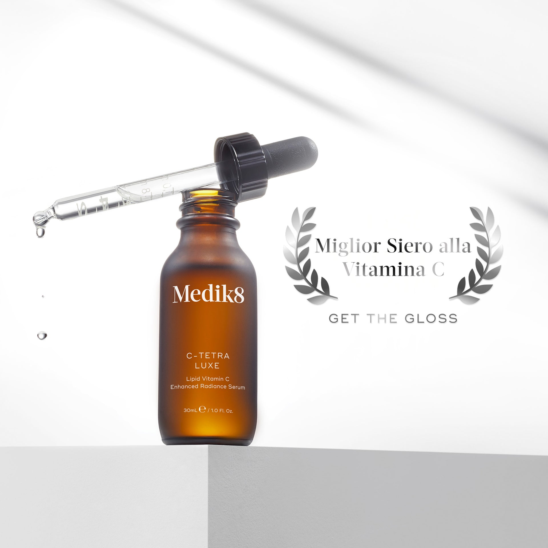 Una boccetta marrone ambrata di siero Medik8 C-Tetra Luxe su un piedistallo grigio chiaro. La pipetta contagocce è appoggiata sopra la boccetta aperta, con gocce di siero che cadono. Sulla destra, un logo a forma di corona d'alloro d'argento riporta la scritta 