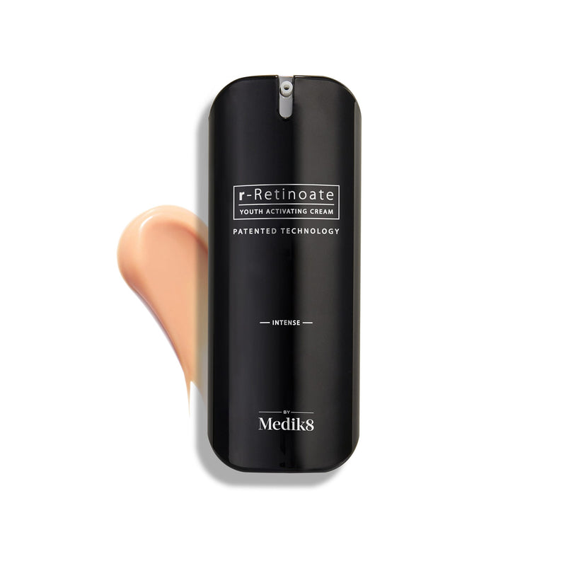 Flacone di Medik8 r-Retinoate Intense Youth Activating Cream esposto su uno sfondo bianco, con una campionatura della crema color pesca visibile sullo sfondo.