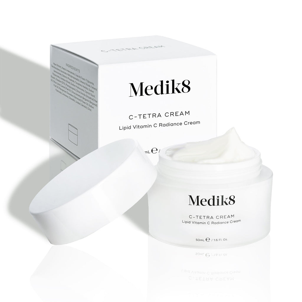 Barattolo di crema Medik8 C-Tetra e la sua confezione in cartone bianco esposti su uno sfondo bianco pulito con ombre morbide. Il barattolo aperto in vetro satinato bianco presenta il logo Medik8 ed è etichettato come C-TETRA CREAM Lipid Vitamin C Radiance Cream da 50 mL.