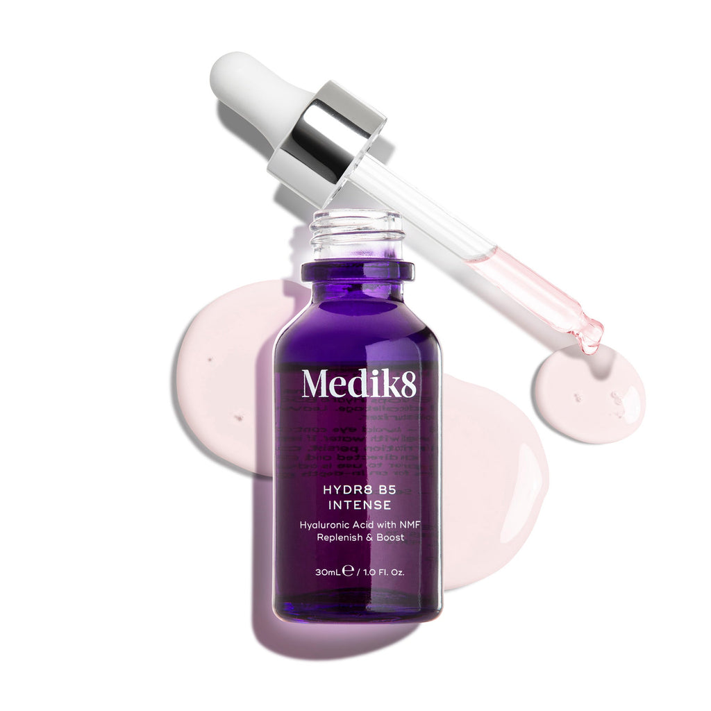 Flacone in vetro di Medik8 Hydr8 B5 Intense di colore viola intenso, mostrato con pipetta aperta appoggiata sopra gocce di siero rosa chiaro su uno sfondo bianco pulito. Il testo evidenzia la formula con Acido Ialuronico e NMF (Fattori Naturali di Idratazione) per Reidratare e Rinforzare, da 30 mL.