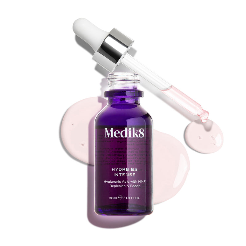 Flacone in vetro di Medik8 Hydr8 B5 Intense di colore viola intenso, mostrato con pipetta aperta appoggiata sopra gocce di siero rosa chiaro su uno sfondo bianco pulito. Il testo evidenzia la formula con Acido Ialuronico e NMF (Fattori Naturali di Idratazione) per Reidratare e Rinforzare, da 30 mL.