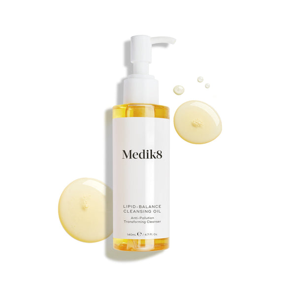 Il flacone di Medik8 Lipid-Balance Cleansing Oil è posizionato su uno sfondo bianco. Gocce di olio dorato circondano il flacone, evidenziandone la texture ricca e fluida.-18