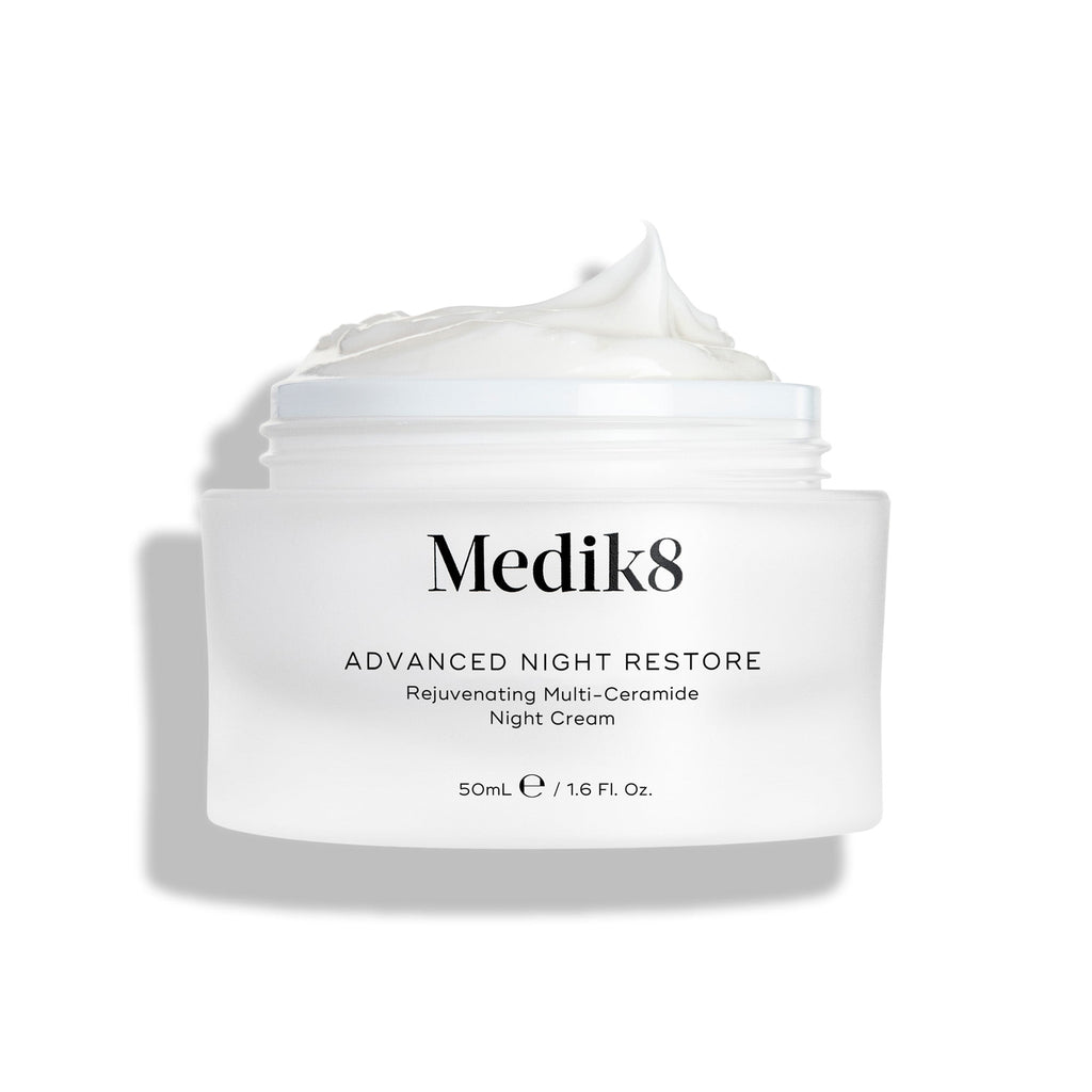 Il barattolo di Medik8 Advanced Night Restore è mostrato aperto con una ricca punta di crema bianca sulla superficie, su uno sfondo bianco pulito che ne evidenzia la texture liscia e rigenerante. Il testo nero sul barattolo riporta Rejuvenating Multi-Ceramide Night Cream.