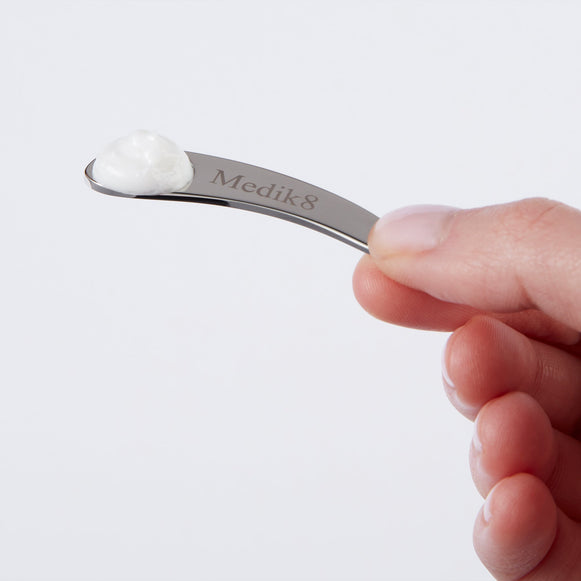 Eine Hand hält einen kleinen, gebogenen silbernen Metallspatel mit dem eingravierten „Medik8“-Logo. Auf der Spitze des Spatels ist ein kleiner Klecks weißer Creme zu sehen.-hover-3