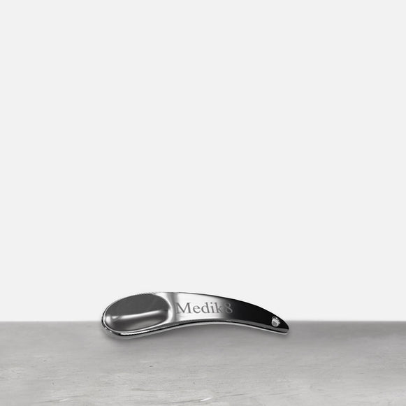 Auf einer grauen Fläche liegt ein silberner Metallspatel. Auf dem Griff des gebogenen Spatels ist das „Medik8“-Logo eingraviert.-3